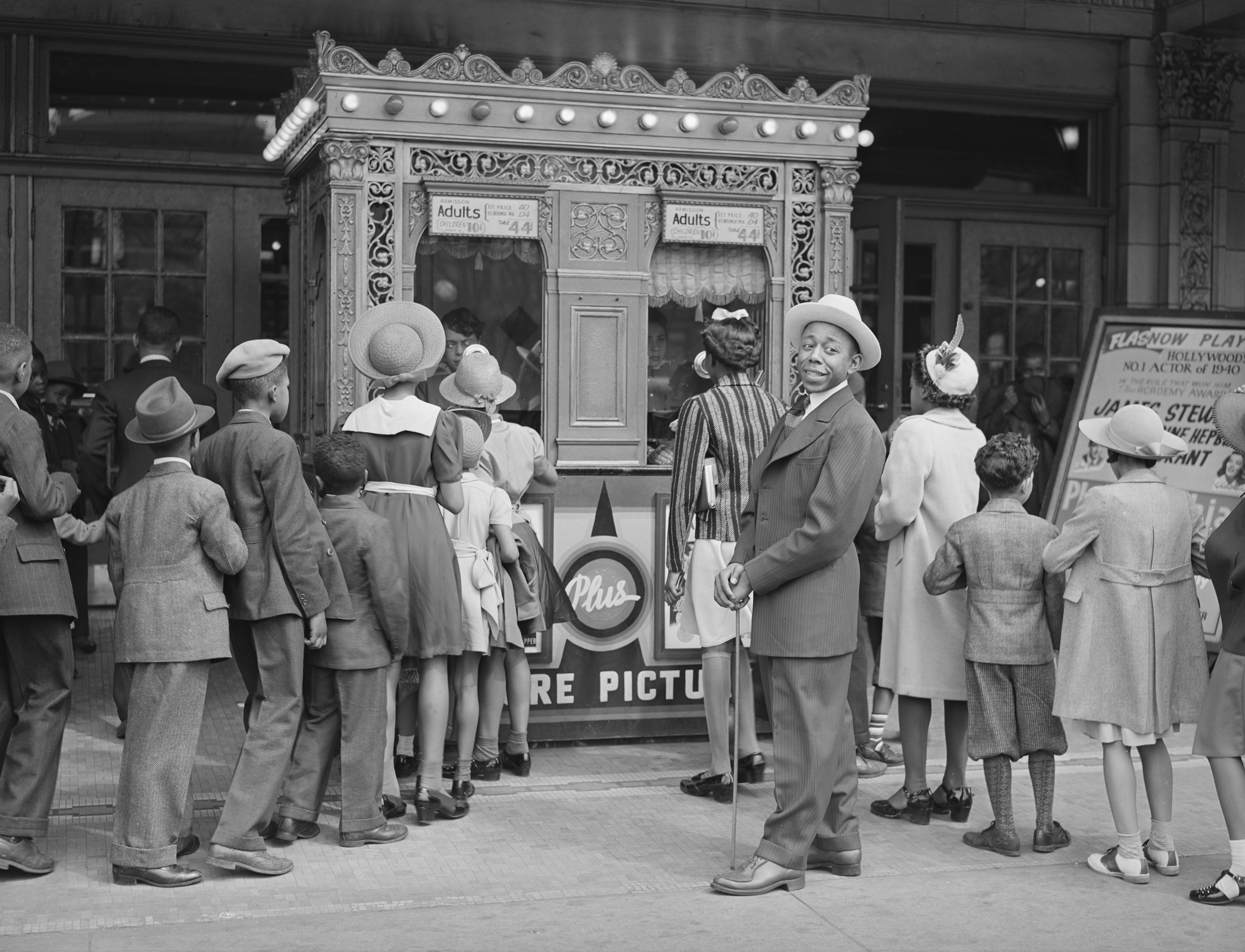 Black-and-white photograph from 1941 showing two lines forming outside a theater to purchase tickets to a movie. A young Black man in a suit and holding a cane looks toward the camera and smiles.